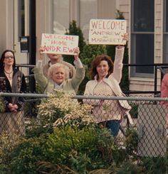 several people holding up signs in front of a fence with bushes and shrubs behind them