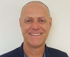 a man in a blue shirt and black blazer smiles at the camera while standing against a white wall