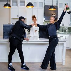 two people standing in front of a kitchen counter holding wine bottles and posing for the camera