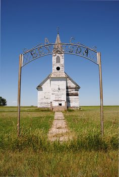 an old white church sits in the middle of a field with a metal arch over it