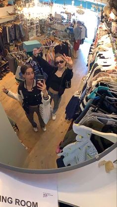 two women standing in the middle of a clothing store looking at their cell phones and taking pictures