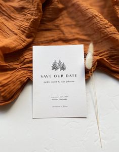 a save the date card sitting on top of an orange cloth with a white feather sticking out of it