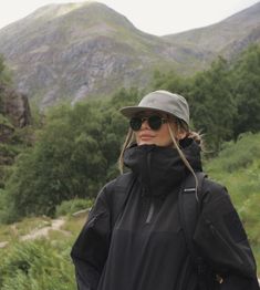 a woman wearing sunglasses and a hat standing in front of a mountain with trees on it