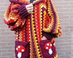 a woman wearing a crocheted jacket with mushrooms on the front and sides, standing in front of a brick wall