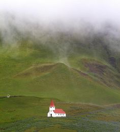 a small white church with a red roof on a green hill covered in fog and clouds