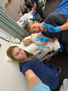 two women in blue gloves are sitting on a hospital bed and one woman is pointing at the camera