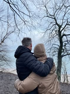 two people hugging each other in front of a tree and the water behind them with no leaves on it