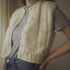 a woman wearing a white knitted vest with holes in the front and back, standing next to a window