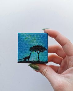 a hand holding up a small painting of a tree and giraffe in the distance