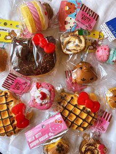 many different kinds of donuts and pastries on a white tablecloth with plastic wrappers