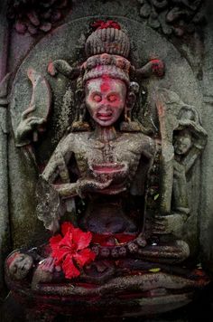 a statue with red paint on it sitting in front of a stone wall next to a flower