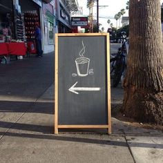 a chalkboard sign with a coffee cup drawn on it next to a tree and sidewalk