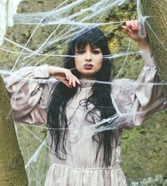 a woman with long black hair wearing a white dress standing next to a tree