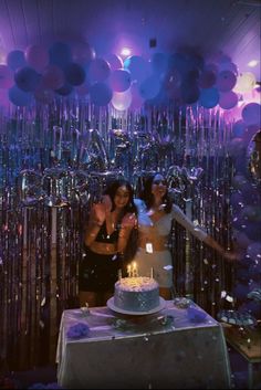 two women standing in front of a cake with candles on it and balloons behind them