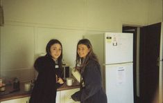 two young women standing in a kitchen next to refrigerators and counter top with food on it