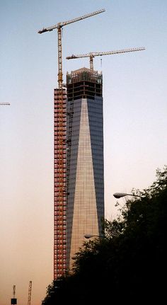 a tall building under construction with cranes on it's side and trees in the foreground