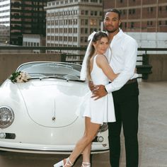 a man and woman standing next to a white car in front of some tall buildings