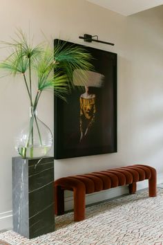 a painting hangs on the wall next to a bench and vase with a plant in it