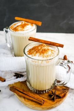 two mugs filled with hot chocolate and cinnamon on a wooden board next to some cinnamon sticks