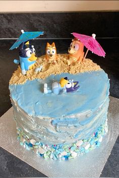 a blue frosted cake with cartoon characters on top and an umbrella in the sand