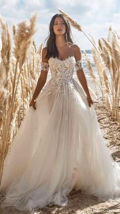 a woman in a white wedding dress standing on the beach with tall grass behind her