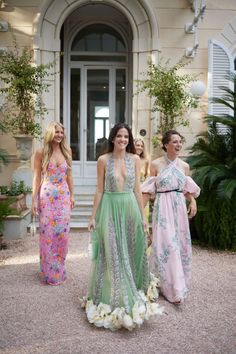 three women standing in front of a building with flowers on the ground and one woman wearing a dress