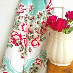 a white vase filled with red flowers sitting on top of a wooden table next to a curtain