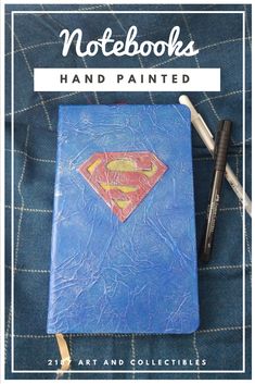 notebooks hand painted with superman logo on blue leather cover and black pen laying next to it