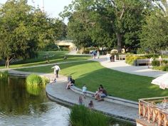 people sitting on the edge of a pond in a park with grass and trees around it