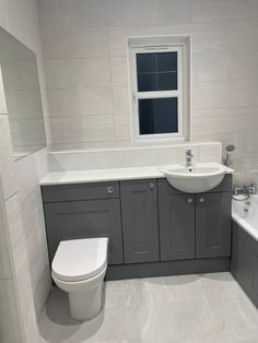 a white toilet sitting next to a bath tub under a window in a bathroom with gray cabinets