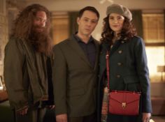 two men and a woman standing next to each other in a living room with a red purse