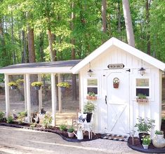 a small white shed sitting in the middle of a forest with potted plants on it