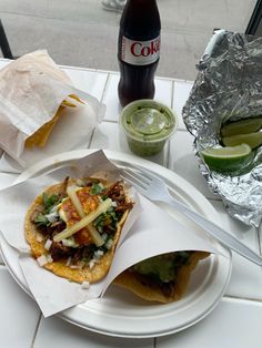 two tacos on a plate next to a bottle of soda and some limeade