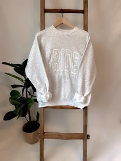 a white sweatshirt with the word bride on it sitting on a wooden chair next to a potted plant