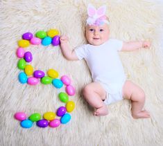 a baby laying on top of a fluffy white blanket next to some candy eggs and an egg shaped letter