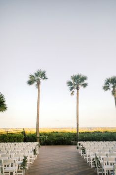 rows of white chairs sitting on top of a wooden floor next to palm trees in front of a field