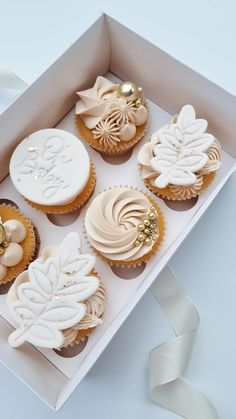 four decorated cupcakes in a box with white ribbon on the side and gold decorations