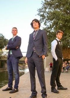 three men in suits standing next to each other on a sidewalk near water and trees