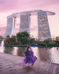 a woman in a purple dress is standing on a dock near some water and tall buildings