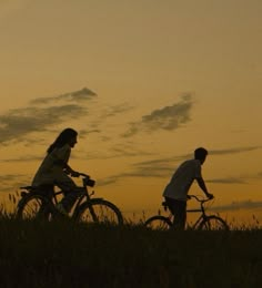 two people riding bikes in the grass at sunset