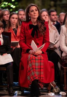 a woman in a red coat and plaid skirt sitting on a chair with other people