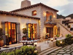 an outdoor patio with tables and chairs next to a stone building at dusk, surrounded by greenery