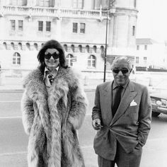 an old black and white photo of two people in fur coats on the street with cars behind them