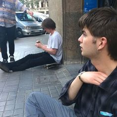 two young men are sitting on the sidewalk and one is using his cell phone while another man stands behind them