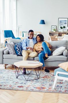 a man and woman sitting on a gray couch in a living room with blue accents