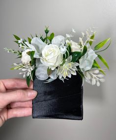 a hand holding a black box with white flowers in it and greenery on top