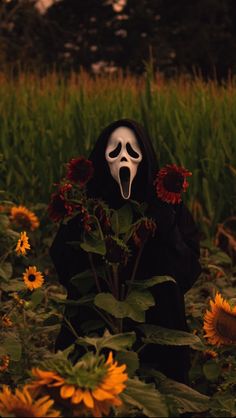 a person in a black mask with sunflowers and grass behind them is holding a white face