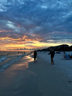 people walking on the beach at sunset with clouds in the sky and water behind them