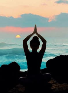 a woman sitting on top of a rock next to the ocean with her hands in the air