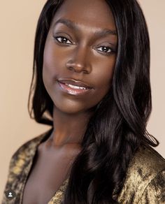 a woman with long dark hair wearing a gold jacket and posing for a photo in front of a beige background